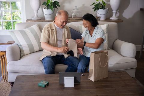 Asian senior couple unwrapping beige garment on sofa while video calling on tablet