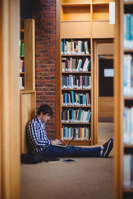 Young adult student sitting on floor in college library, using laptop for studying. Surrounded by bookshelves, focused on work. Ideal for educational content, technology in education, academic research, and student life themes.