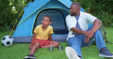 African american father and son camping and bonding in backyard on grass with blue tent