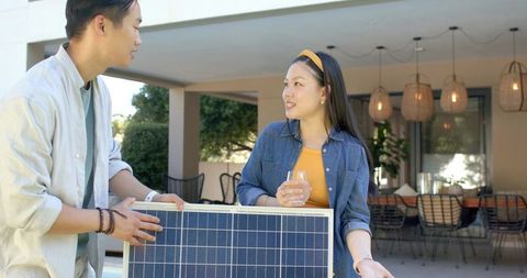Asian couple holding portable solar panel on covered patio while enjoying drink and conversation