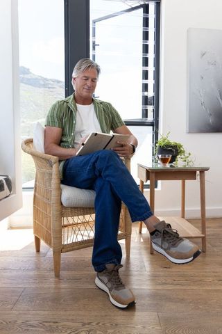 Senior man relaxing with tablet in sunlit wicker chair by window beside wooden side table
