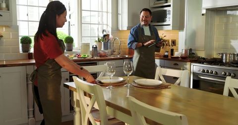 Middle-aged Asian man and partner arranging farmhouse table, cooking together in bright kitchen