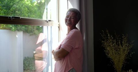 Senior woman smiling by sunlit window with reflection, relaxed home lifestyle portrait