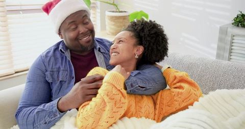 Couple sharing cozy holiday cuddle on sunlit couch with chunky blanket and Santa hat
