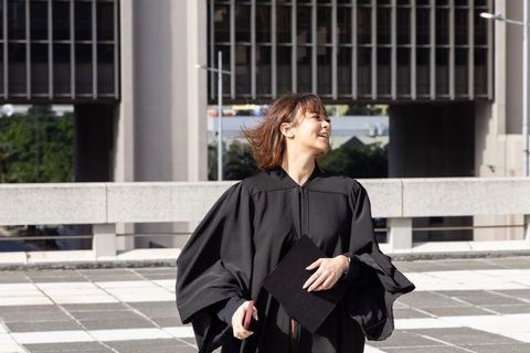 Asian woman graduating on urban terrace celebrating holding mortarboard and smartphone