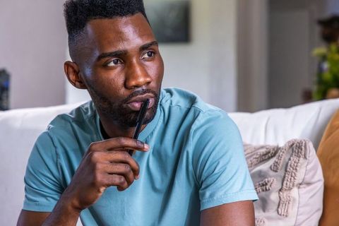 Contemplative african american man holding pen on sofa, thinking in modern living room