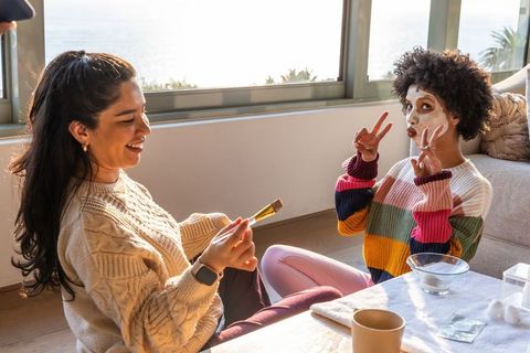 Friends enjoying coastal self-care ritual applying face masks and laughing in sunlit home
