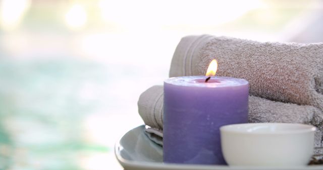 Lit lavender candle and neatly stacked towels on a plate, creating a perfect spa-like atmosphere. Ideal for conveying relaxation, wellness, self-care, aromatherapy, and serene environments. Perfect for spa advertisements, relaxation blogs, wellness websites, and self-care promotion materials.