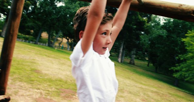 Young boy in white shirt hanging on monkey bars in park, enjoying outdoor activity. Trees and grass provide the natural background. Ideal for use in articles on childhood development, active lifestyle, outdoor activities for kids, or park recreational areas.