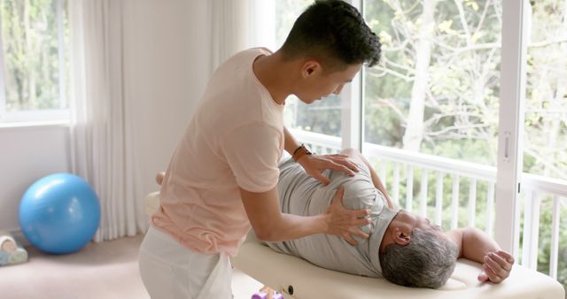 A physical therapist is treating the shoulder of a senior man, providing therapy in a bright, comfortable room. This could be used in materials related to physical therapy, healthcare services, senior care, or wellness programs. The file might be ideal for websites or promotional content focused on pain management, rehabilitation, or medical treatments.