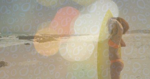 Surfer standing with yellow surfboard on sunlit beach, bokeh double exposure lens flare