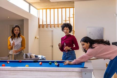 Diverse women playing pool in modern home with blue-felt table and natural skylight