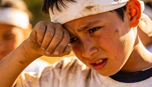 Dirt-smudged boy wiping eye during outdoor game with white headband and dusty uniform