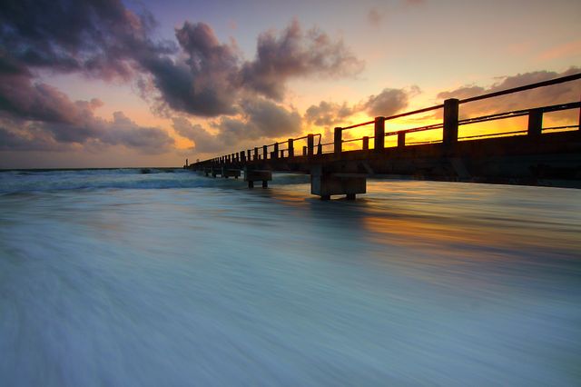 This serene scene shows an ocean pier at sunrise with gentle waves and a colorful sky filled with clouds. Ideal for travel advertisements, vacation brochures, desktop wallpapers, and relaxation promotions. Perfect for depicting tranquil and peaceful coastal environments.
