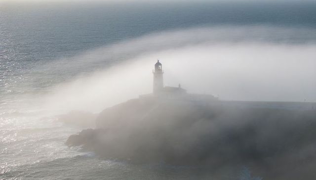 Lighthouse emerging from coastal fog on rugged headland, waves crashing, atmospheric seascape