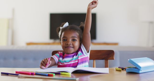 African American girl with raised hand sitting at wooden table, smiling confidently while doing homework at home. Background includes bookshelf and furniture, emphasizing cozy and conducive home learning environment. Perfect for educational articles, tutoring services, homeschooling resources, and child development content.