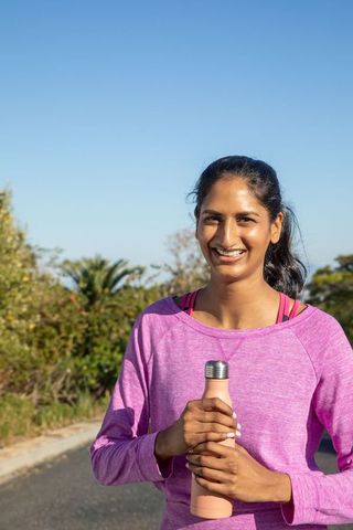 Indian woman standing outdoors holding water bottle smiling after morning run in pink top