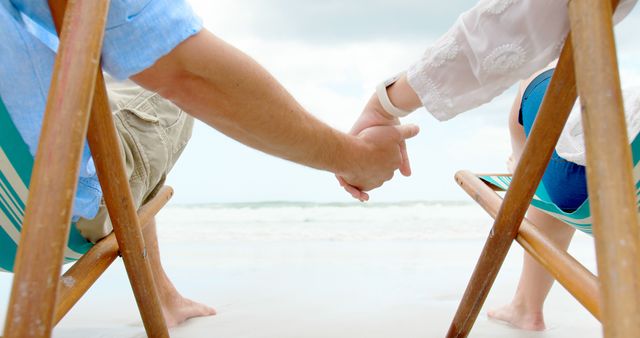 Midsection of caucasian couple sitting in deckchairs holding hands on beach. Relationship, togetherness, relaxation, summer, leisure, vacation and lifestyle, unaltered.
