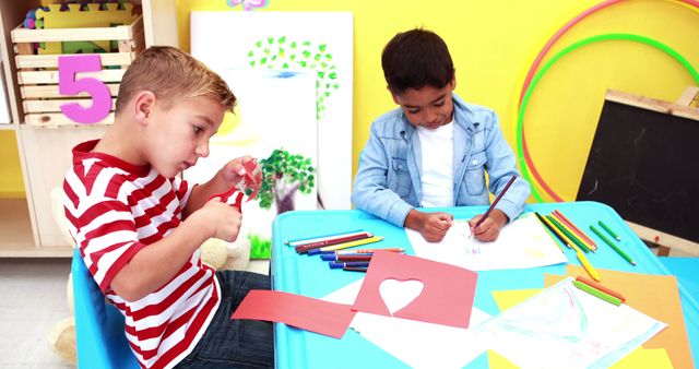 Two children at a classroom table engage in art and craft activities. One child draws with colored pencils, while another cuts paper for a craft project. The background is bright and colorful, promoting creativity and learning. Ideal for use in educational materials, classroom activity guides, children's care brochures, and resources on childhood education and development.