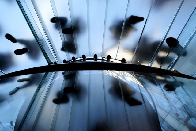 Underside view of people ascending a glass staircase. Evidence of modern architectural trends with emphasis on transparency and geometric lines. This shot is ideal for use in advertisements for architectural firms, urban-related design features, or technology products showcasing modernity and innovation. Suitable for promoting architectural tours or illustrating concepts in business materials.