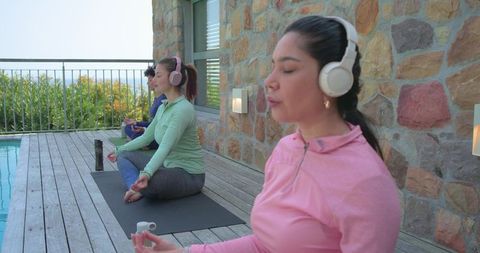 Diverse women meditating with headphones on poolside wooden deck for mindfulness