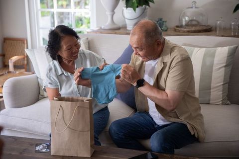 Asian grandparents discovering baby onesie surprise on cozy living room sofa