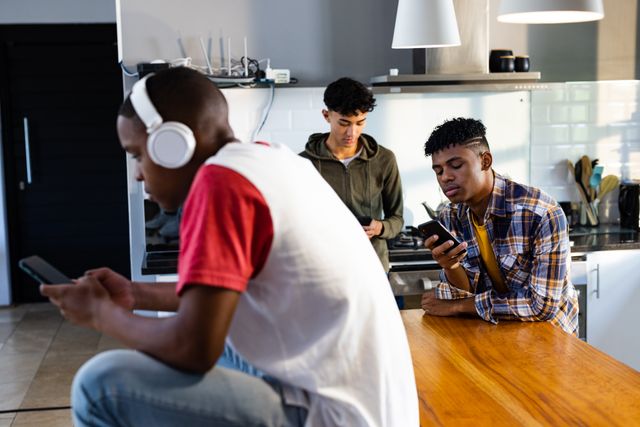 Diverse teenager male friends using smartphones in kitchen. Hanging out with friends and spending quality time together concept.