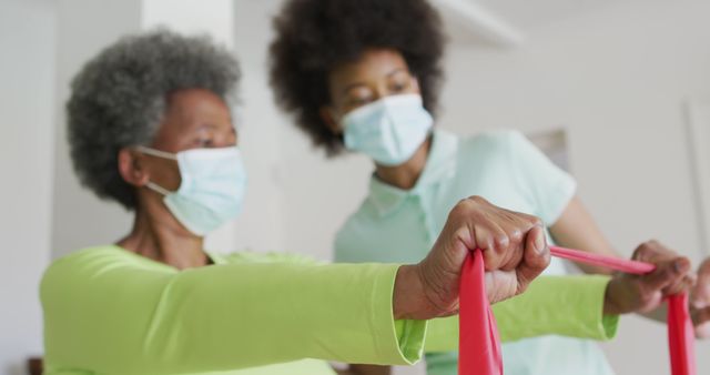 Two women wearing masks engaging in a physiotherapy session. The senior woman is using a resistance band under the supervision of a physiotherapist. Useful for promoting health services, illustrating rehabilitation exercises, and senior wellness.