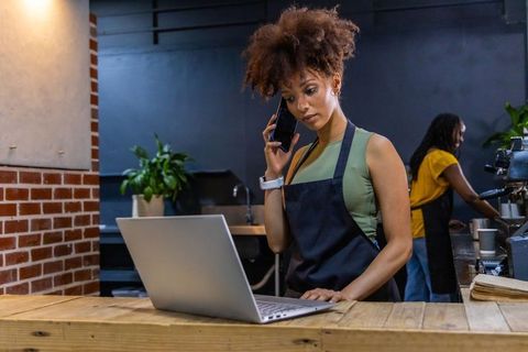 African american barista managing cafe orders with laptop and phone at wooden counter