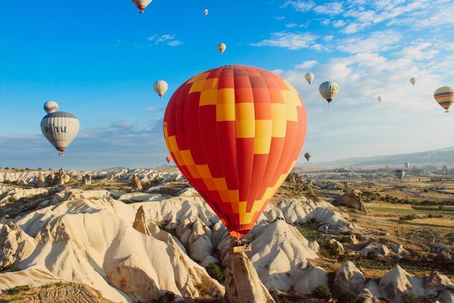 Multiple hot air balloons are flying over the unique rocky terrain of Cappadocia, Turkey. The bright complementary colors contrast with the golden sunlight, creating a picturesque and vibrant scene. Ideal for promoting travel agencies, adventure tours, Turkish tourism, and aerial photography.