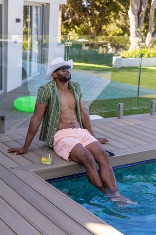 Mature african american man relaxing on pool deck with drink, hat and sunglasses