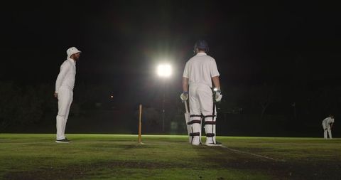 Floodlit night cricket match with helmeted batsman standing at crease and umpire watching