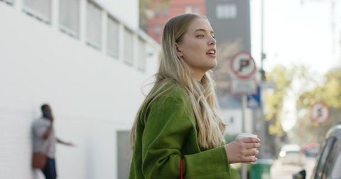 Blonde Woman Standing on Urban Sidewalk Wearing Green Coat Holding Coffee Cup, City Lifestyle