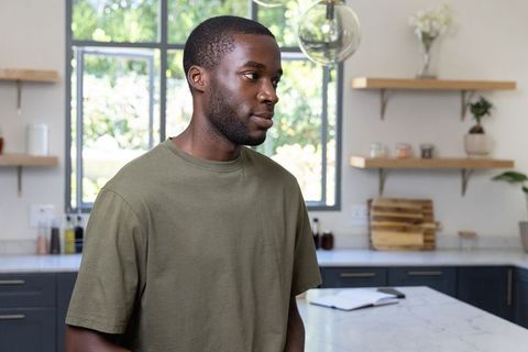 Thoughtful african american man standing at marble island holding planner in sunlit kitchen