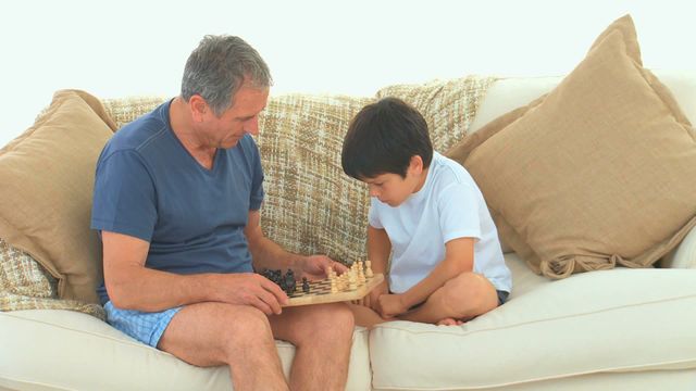 Grandfather and grandson are engaged in a chess game on a comfortable sofa, creating a scene of family bonding and intellectual exchange. This is ideal for visuals related to family relationships, upbringing, educational activities, and leisure at home. Perfect for articles or advertisements highlighting nurturing family dynamics, teaching moments, or the benefits of strategy games.