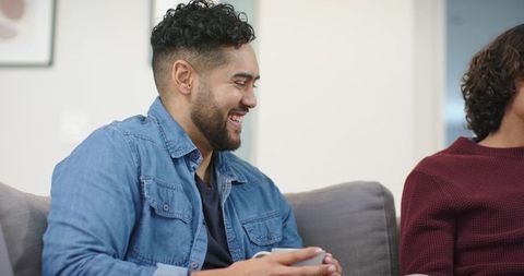 Friends laughing on couch sharing coffee, man in denim jacket holding mug in cozy conversation