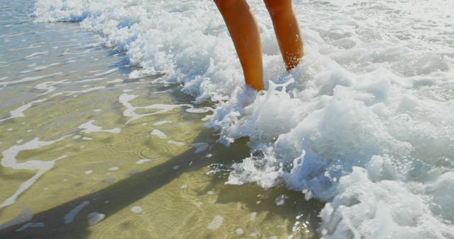 Feet and legs wading in gentle ocean waves on a sandy beach. Suitable for themes of relaxation, summer vacations, seaside activities, and nature. Ideal for travel advertisements, beach resort promotions, and wellness campaigns.