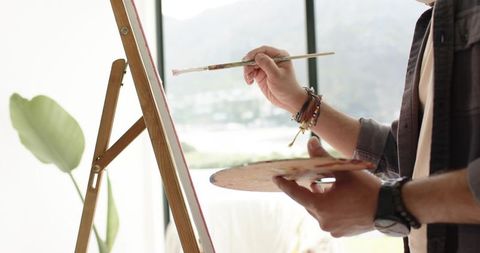 Indian man painting on stretched canvas in sunlit studio by window holding palette and brush
