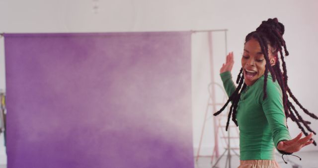 African American woman dancing in a studio setting with a vibrant purple backdrop. Her long dreads are in motion, and she is expressing joy and excitement. The image exudes energy, happiness, and freedom, making it ideal for use in advertisements or campaigns promoting vitality, creativity, and fun. Perfect for lifestyle blogs, social media posts, or fitness promotions.