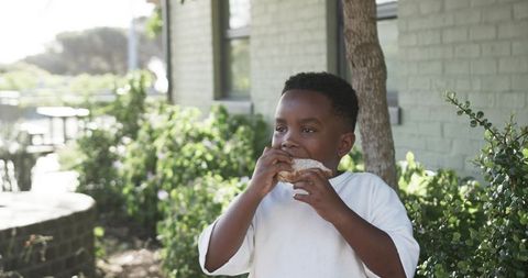 Young boy enjoying sandwich outdoors in sunny garden by brick house