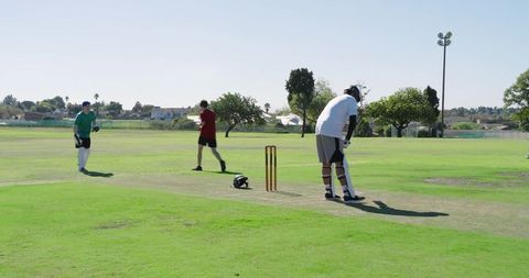Amateur cricket players competing on sunlit park pitch, bowler running while batsman sprinting