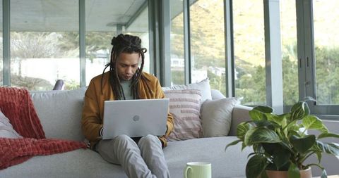 African american man working remotely on laptop on modern sofa with plants and large windows
