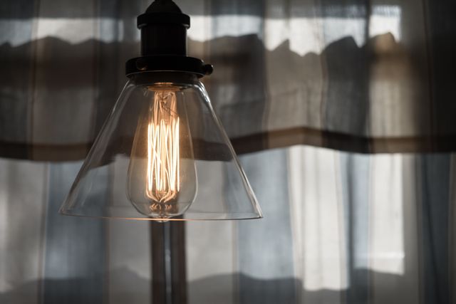 Image depicts a warm glowing vintage Edison light bulb with exposed filament, hanging against sheer, softly lit curtains. Ideal for illustrating articles or blogs about interior design trends, home lighting solutions, or creating a cozy atmosphere. Useful for designers or decorators seeking inspirational decor elements in their portfolios.