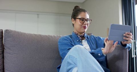 Young woman sitting on sofa using tablet wearing tortoise-frame glasses and silk scarf