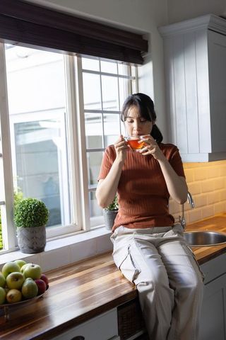 Asian woman sipping tea while sitting on kitchen countertop by sunlit window