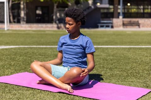 African american boy sitting cross-legged on yoga mat on turf looking thoughtful outdoors