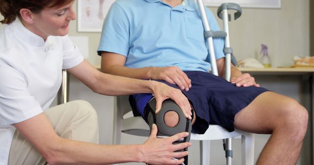 Physical therapist assists patient with knee brace, indicative of medical rehabilitation. Patient sits, holding crutch, conveying recent injury or surgery recovery. Useful for healthcare websites, rehabilitation service promotions, medical education materials, and content related to physical therapy and orthopedic care.
