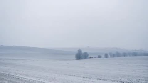 Drone traveling over snow-covered rolling fields with isolated farm and frosty trees