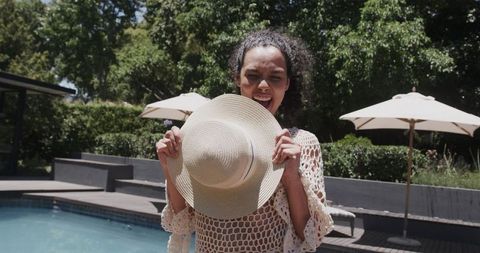 African american woman holding straw hat smiling by backyard pool wearing crochet cover-up