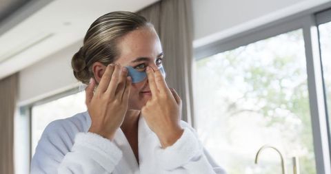 Applying blue hydrogel eye patches in sunlit luxury bathroom, morning skincare ritual